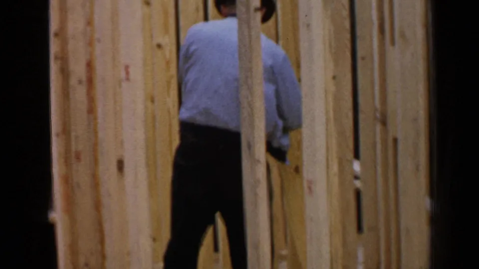 A worker in dark trousers and a light shirt stands among wall studs during a 1957 Birmingham house-framing scene.