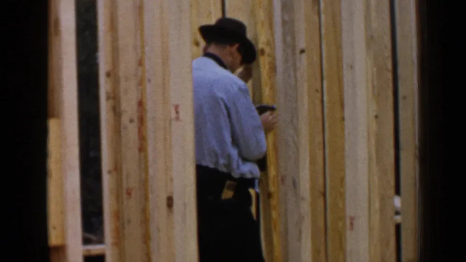 A hat-wearing worker stands close to the wall framing, appearing to inspect or fasten lumber in the Birmingham construction footage.