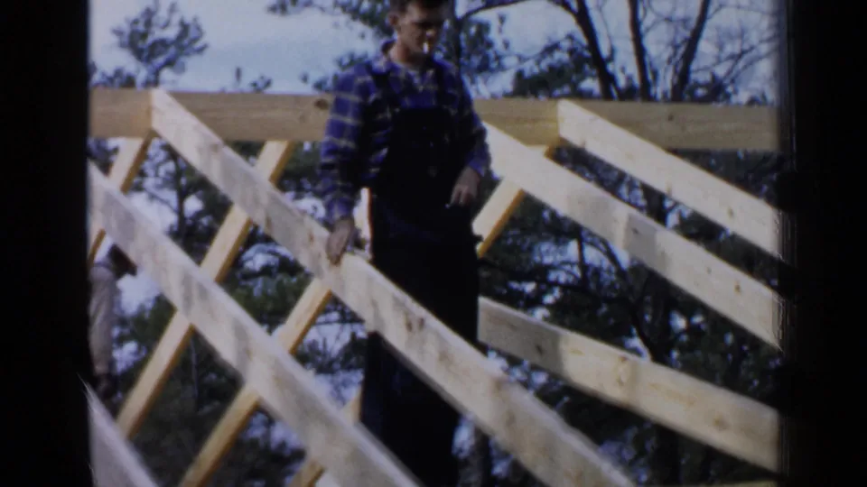 A worker in overalls and a plaid shirt handles lumber among angled roof beams, with trees visible beyond the frame.