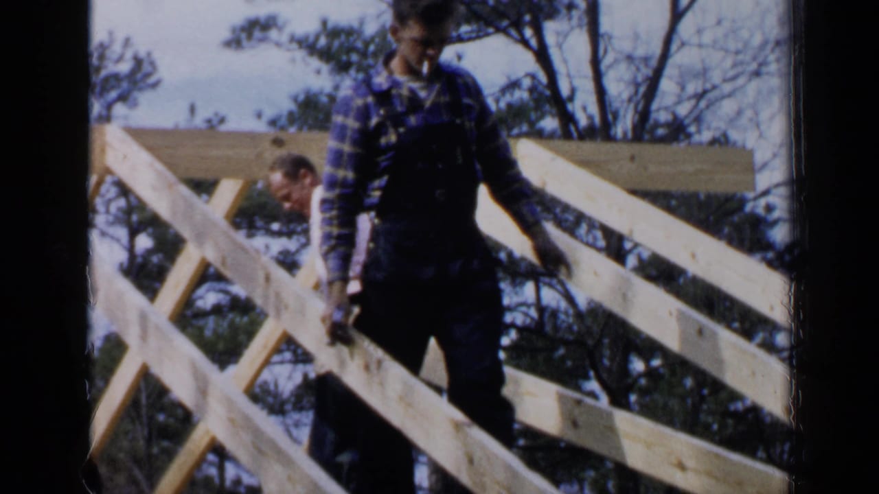 Two men work among pale wooden roof beams outdoors.