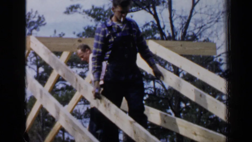 In Birmingham, Alabama, 1957, two adult construction workers stand among diagonal wooden roof framing, with trees and sky behind them.