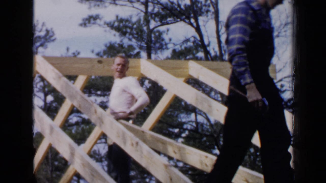 A man in a light shirt stands behind roof framing while another worker passes at right.