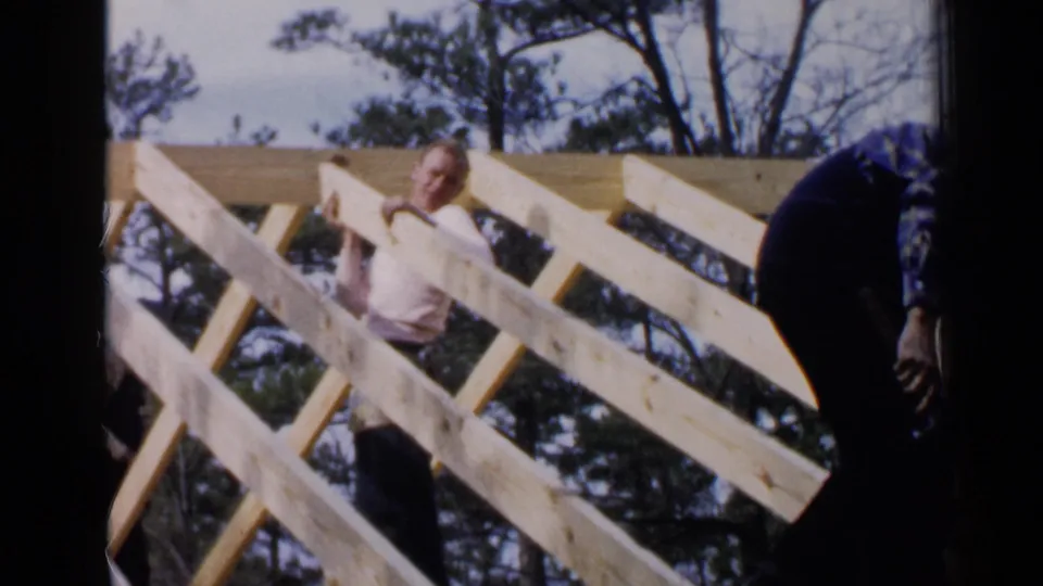 A man in a light long-sleeve shirt handles a piece of lumber within an unfinished roof structure in Birmingham, Alabama, based on the supplied clip metadata.