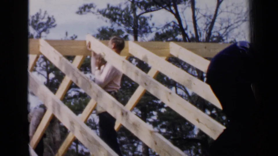A construction worker appears to lift or steady a board inside the diagonal roof frame, with another worker partially visible at the right edge.