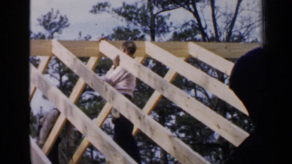 1957 Birmingham home-movie frame showing a man working on a wooden roof frame with pine trees in the background.