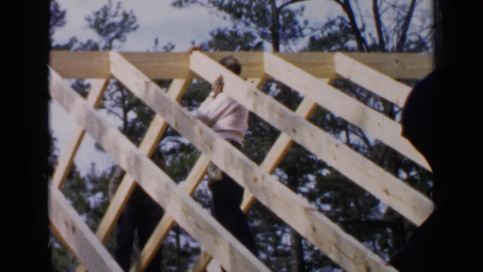 A man works on pale roof rafters in Birmingham, Alabama, while a second figure is silhouetted at the edge of the frame.