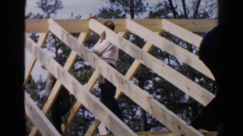 Home-movie view of a worker among exposed roof rafters, with a dark foreground shape partially blocking the construction scene.