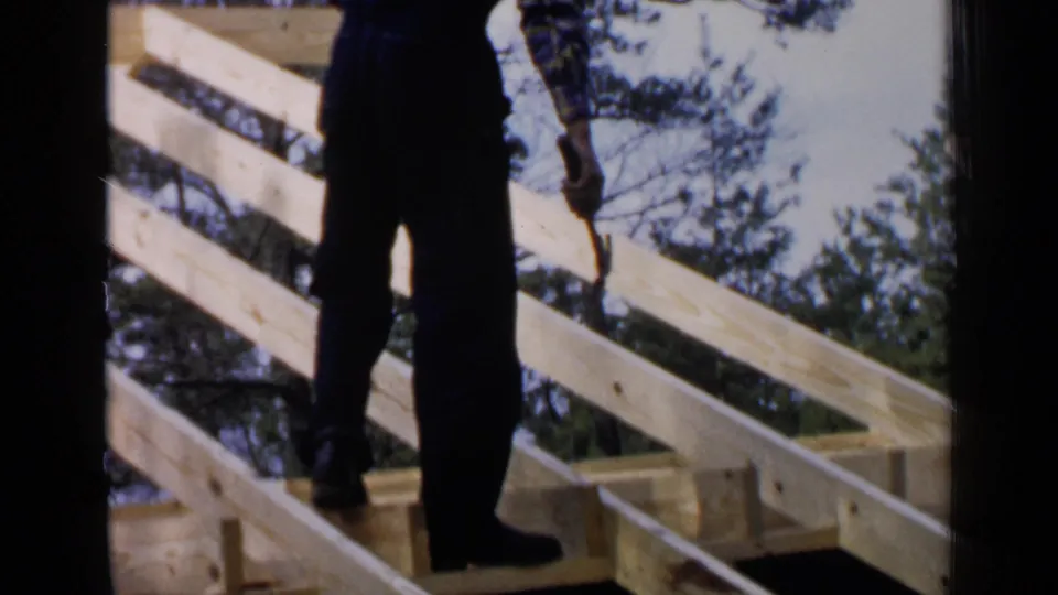 A dark-clothed figure stands on the wooden roof frame during the 1957 Birmingham construction scene.