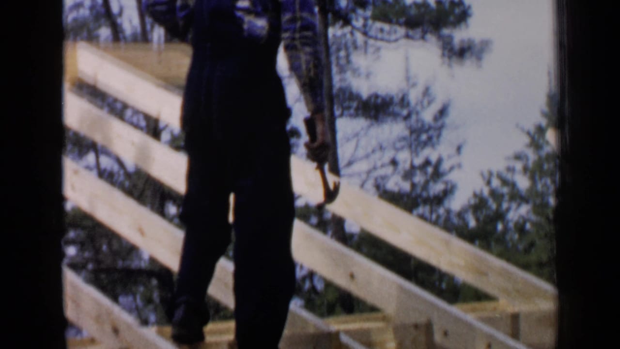 In this 1957 Birmingham, Alabama 8mm frame, a man in dark overalls stands on pale wooden roof framing with a hammer beside diagonal beams and trees.