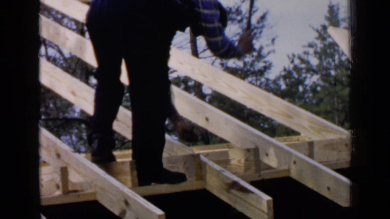 A worker in dark overalls and plaid sleeves moves across exposed wooden beams in the wooded Birmingham, Alabama construction scene from 1957.