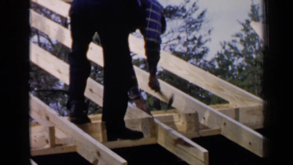 The man bends over the wooden roof frame, placing or striking with a hammer as sunlight catches the pale lumber and trees behind him.