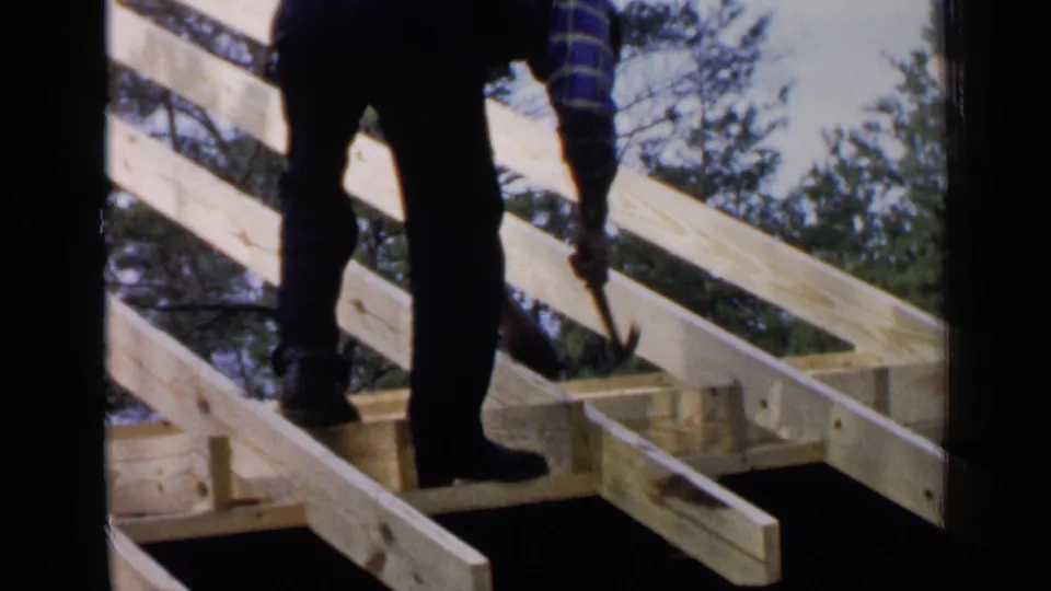 A man in work clothes hammers near the edge of a wooden roof frame in the 1957 Birmingham, Alabama home-movie footage, with trees visible behind the structure.