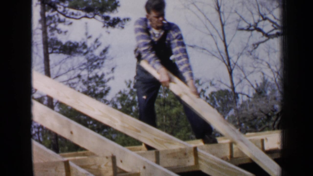 Man in plaid shirt and overalls positions long lumber on a wooden frame outdoors.