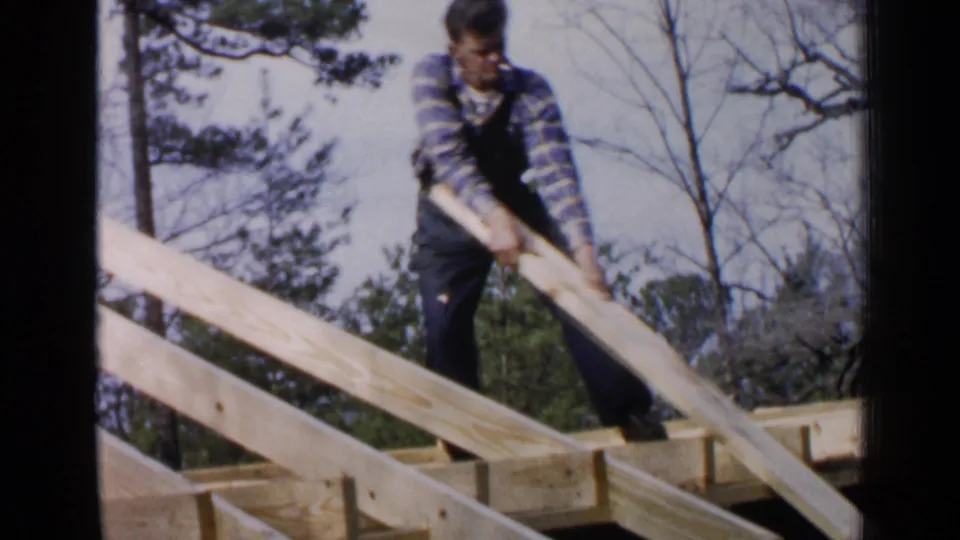 In 1957 Birmingham home-movie footage, a man in plaid and overalls works on roof framing among trees.