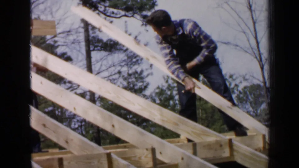 A man bends over pale rafters, shifting lumber on an outdoor wooden frame in Birmingham, Alabama.