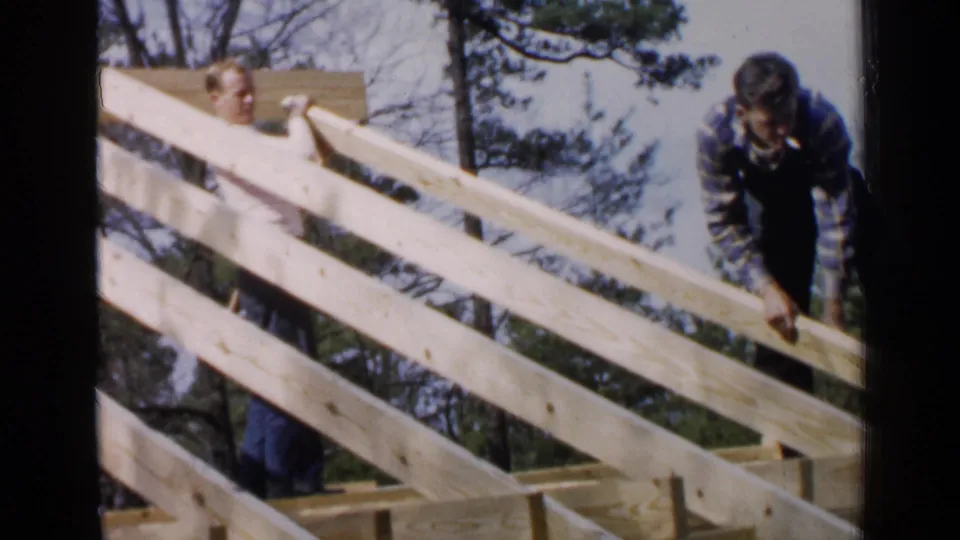 Two men are visible around the partially built frame, one behind the rafters and one bending forward.
