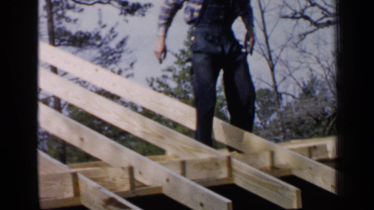 Man standing on a wooden roof frame with rafters and trees behind him.