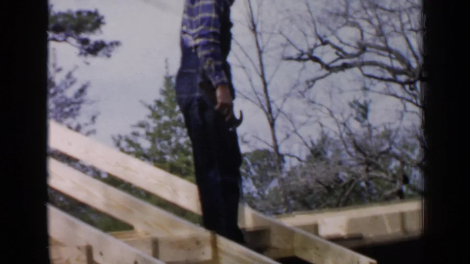 A man appears beside sloped roof rafters in 1957 Birmingham footage, holding a hammer as he works on the wood frame.