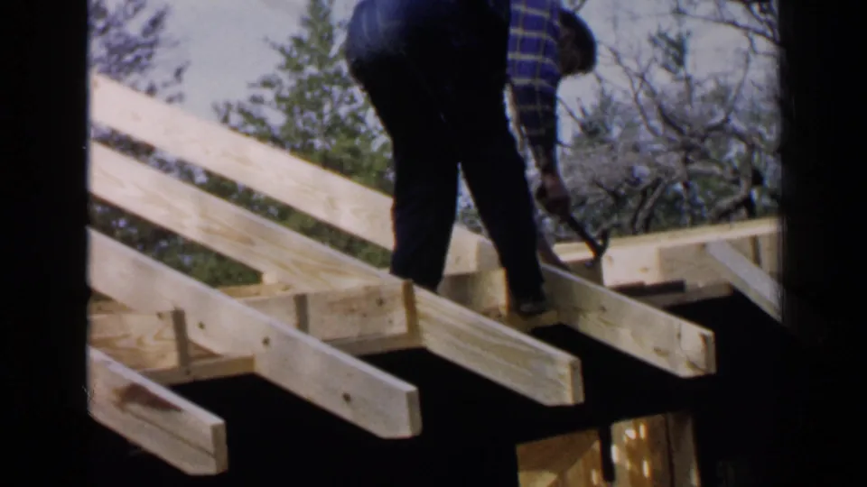 The worker bends over the roof structure, reaching toward exposed rafters in the Birmingham construction scene.