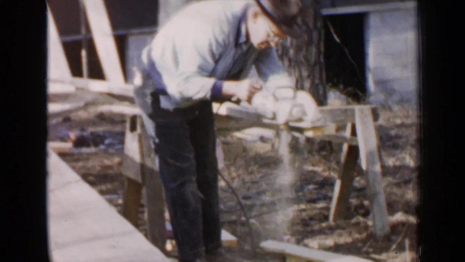 A man cuts lumber with a power saw outdoors near a house wall, continuing the 1957 Birmingham construction work.