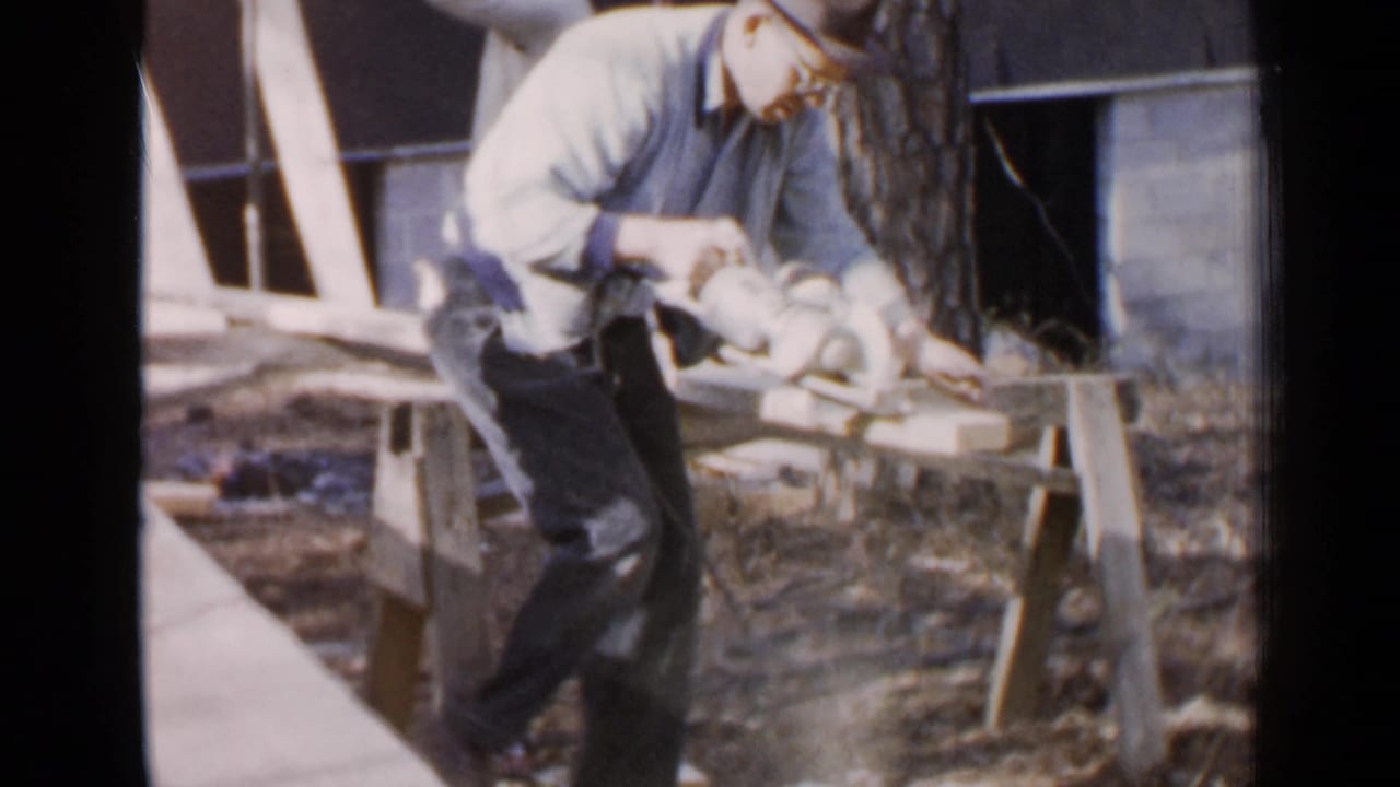 Older man in a fedora uses an electric saw on a plank outdoors.