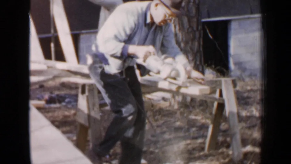 In 1957 Birmingham home-movie footage, an older man in a hat and glasses cuts a plank on a sawhorse with a corded electric saw.