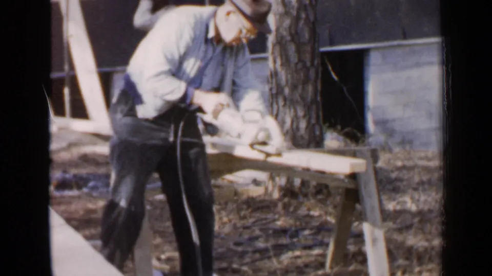 The man leans into the cut, holding a corded electric saw against a board in a Birmingham backyard scene from 1957.