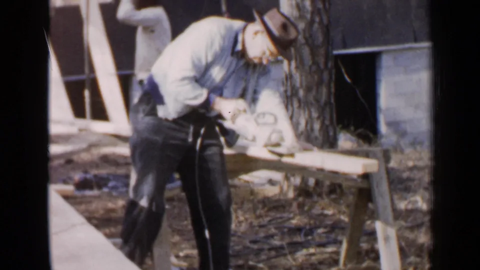 An older man in dark trousers and a light shirt continues cutting lumber with a corded saw in the 1957 Birmingham home movie.