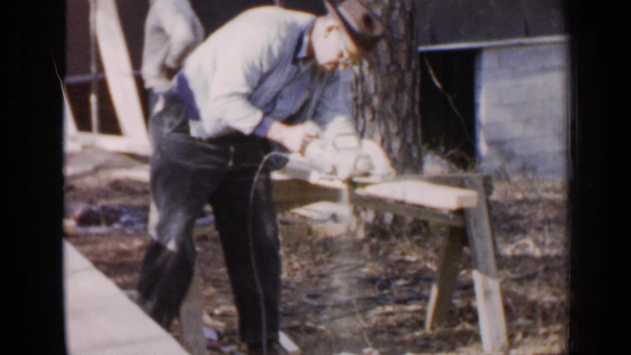 In Birmingham, Alabama, 1957, a man in work clothes and a cowboy hat leans over wooden planks near a tree and brick wall.