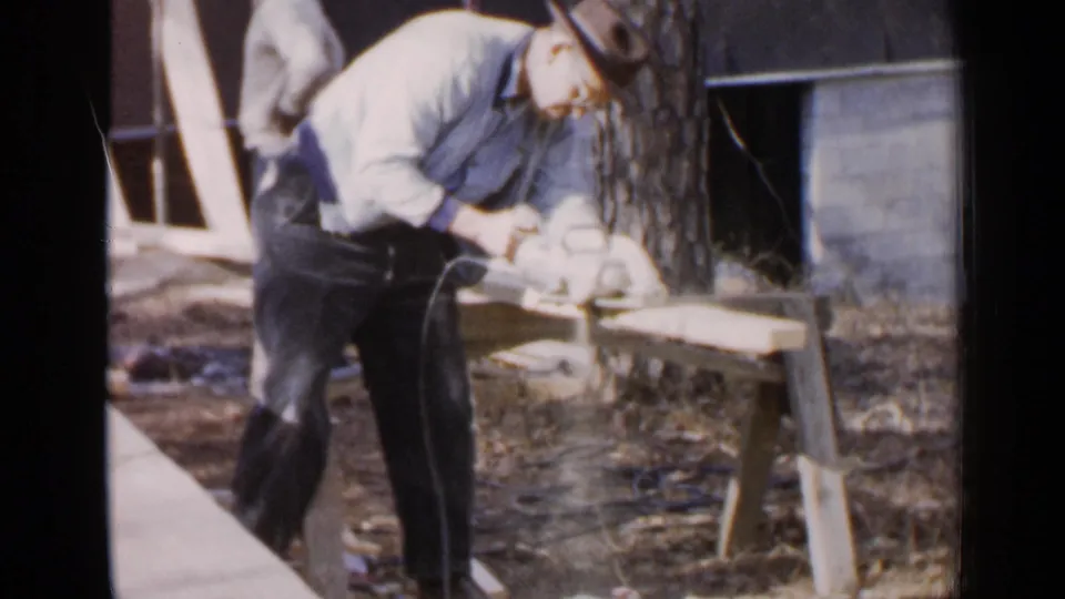 In Birmingham, Alabama, 1957, a man in work clothes and a cowboy hat leans over wooden planks near a tree and brick wall.