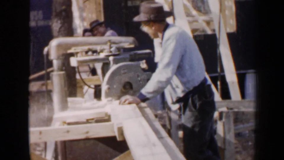 A 1957 Birmingham home-movie frame shows the man beside a circular saw, with lumber, supports, and a brick building behind him.