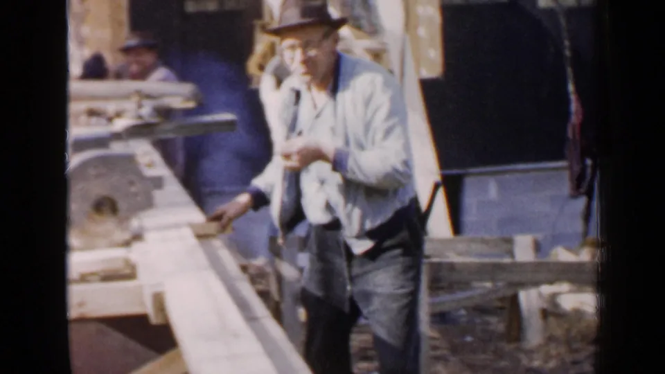The worker briefly faces the camera near the saw and planks, with the Birmingham outdoor construction setting visible behind him.