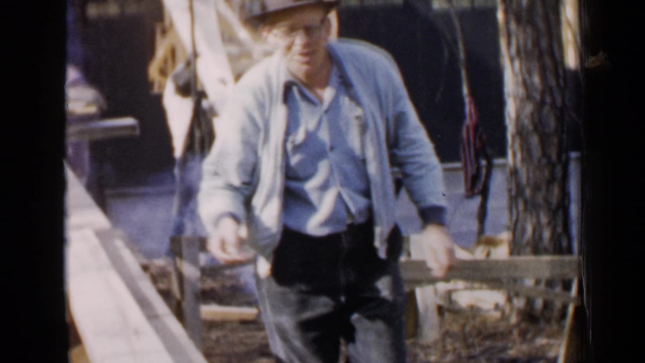 Man in a fedora and glasses walks near an outdoor work area with lumber behind him.