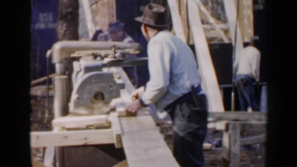 The man positions lumber on an outdoor workbench beside a circular miter saw, with another person visible in the background.