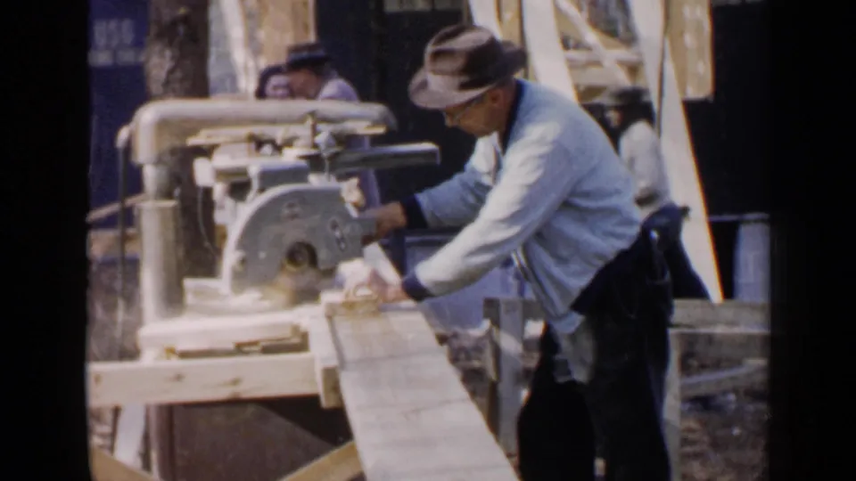 The man works over the miter saw with both hands on the board in the 1957 Birmingham outdoor workshop scene.
