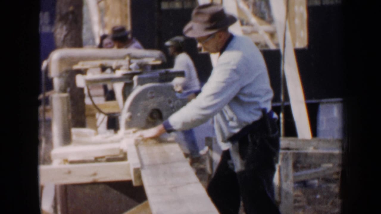 Worker in a fedora guiding a board through a table saw at a construction site.
