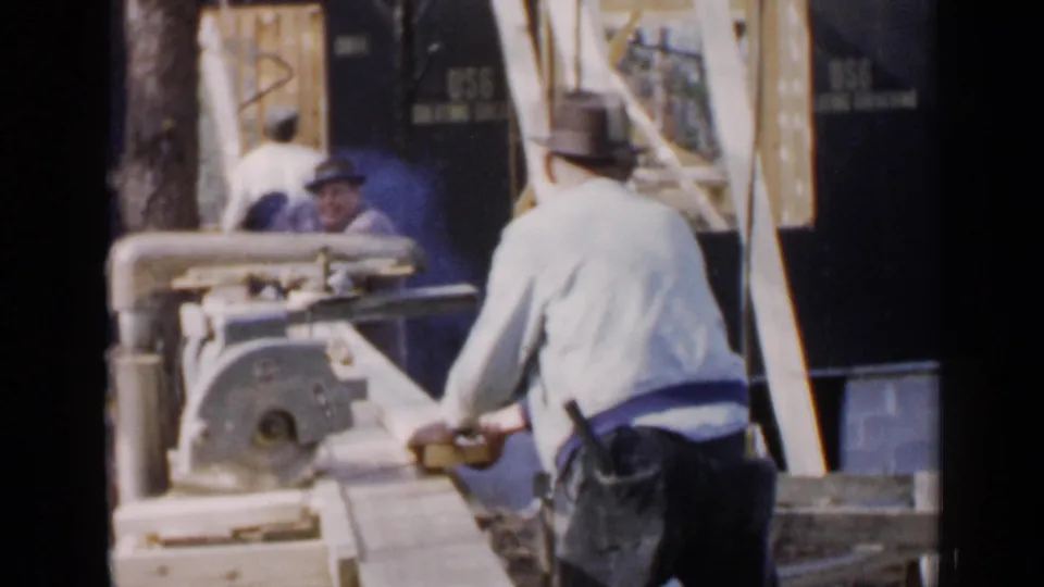 1957 Birmingham construction frame with a worker seen from behind at a table saw, with lumber and framing around him.