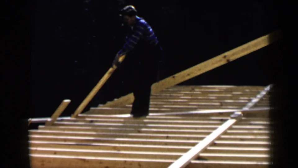 A man in a blue striped shirt works on a sloped wooden roof frame in the Birmingham, Alabama construction footage.