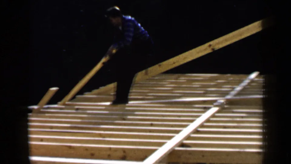 The worker stands on exposed joists and guides a long board across the roof frame in this 1957 Birmingham home-movie scene.