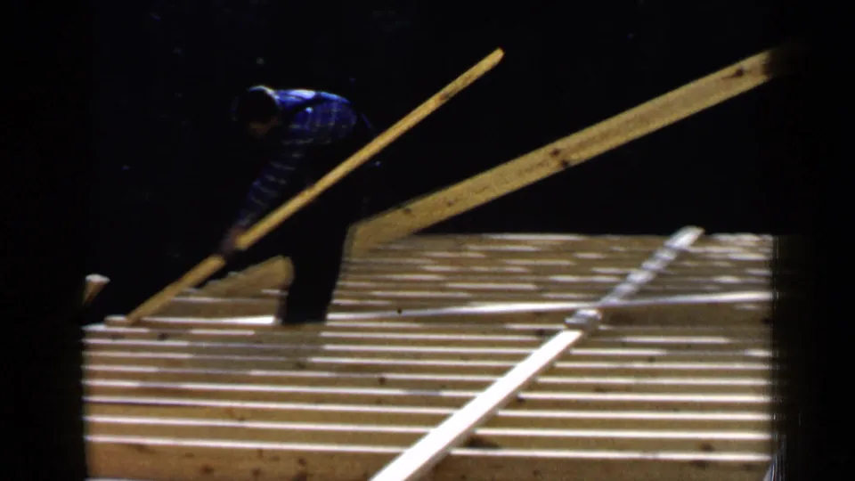 A man bends over the exposed roof frame while handling a long board in the 1957 Birmingham construction footage.