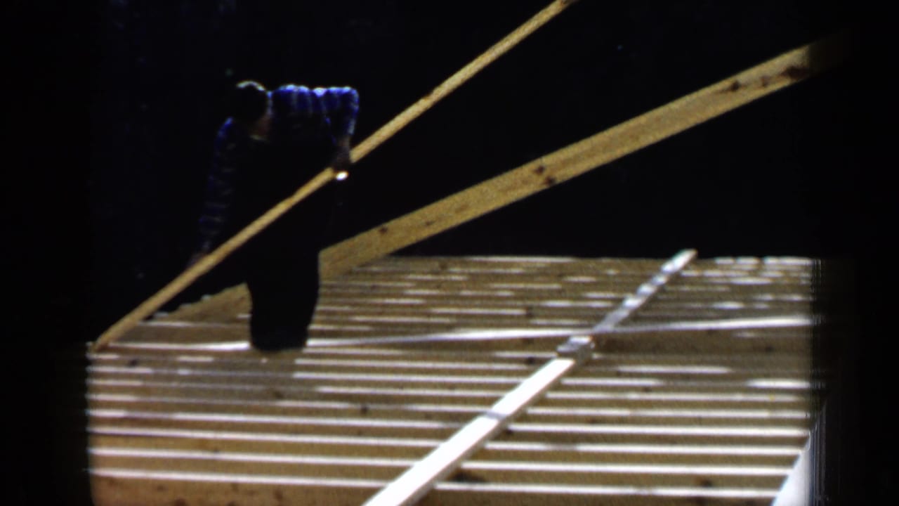Worker carrying a long board above exposed roof framing in bright sunlight.