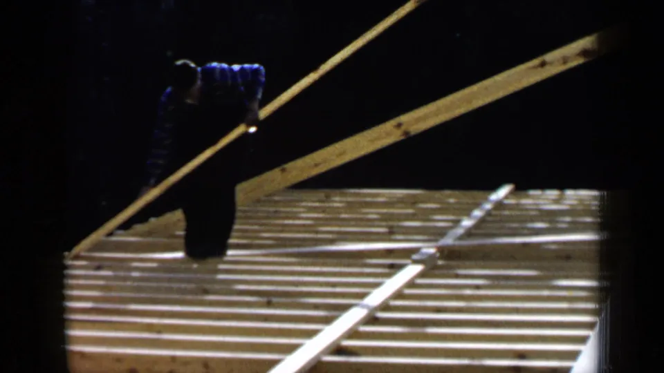 1957 Birmingham footage shows a worker in a striped shirt moving a long board across exposed wooden roof framing.