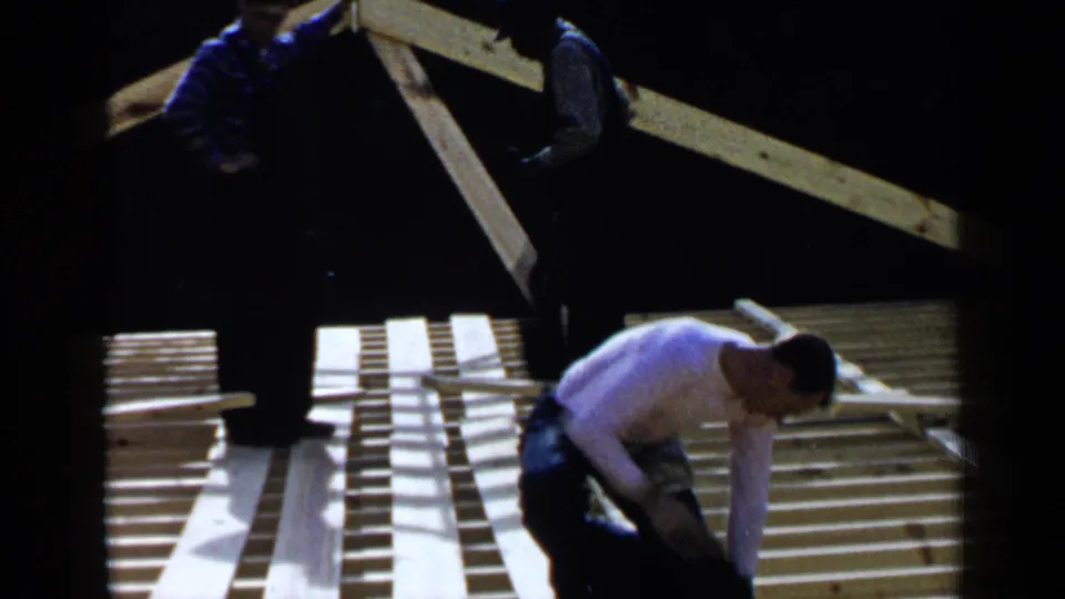 Men on a Birmingham roof frame work among joists and planks, with one man bending close to the lumber.