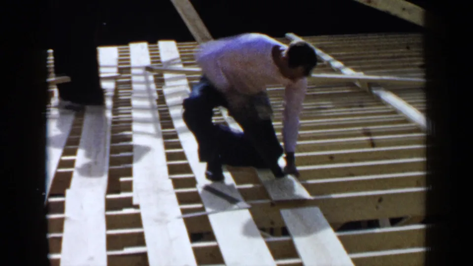 A man in a white shirt kneels on the exposed roof frame, working beside long boards in the Birmingham 1957 footage.