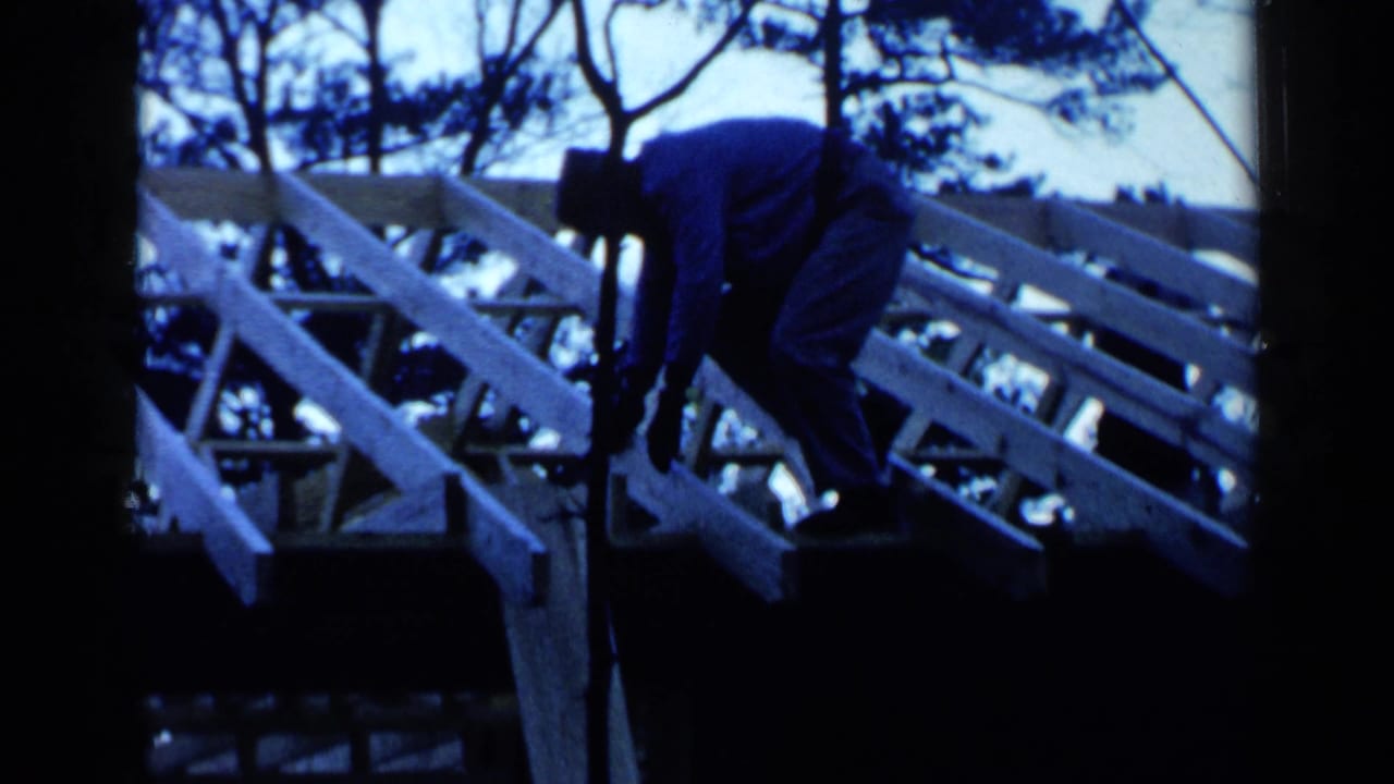 Man in a hat bends over unfinished wooden roof rafters outdoors.