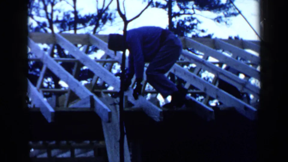 A hat-wearing man works along an unfinished roof frame in Birmingham, Alabama, with pale rafters and trees visible in the background.