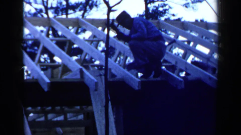 The man crouches on the unfinished roof structure, appearing to work by hand among the rafters in the 1957 Birmingham footage.