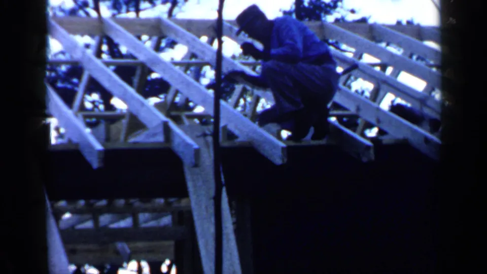 A man in a hat crouches beside exposed wooden rafters, with a vertical support and trees visible in this Birmingham home-movie frame.