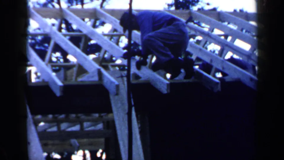 A man in dark clothes and hat bends over the timber roof frame in the 1957 Birmingham home-movie footage.