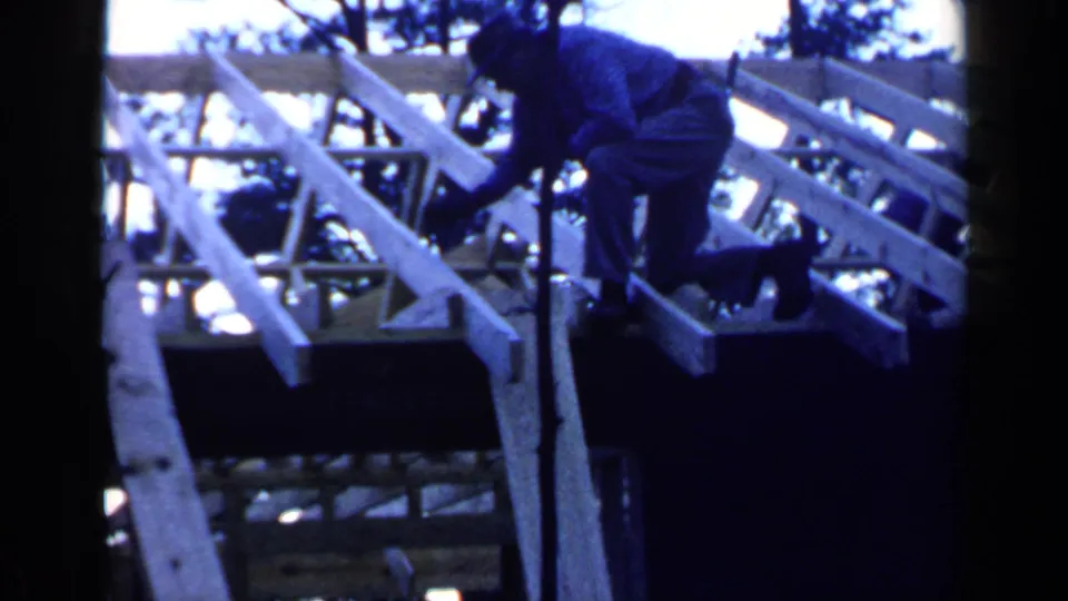 The worker appears to hammer or adjust a beam while crouched on the sloped roof framing in Birmingham, Alabama, in 1957.